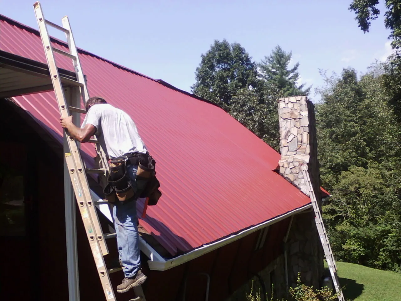 Roofer on a ladder beside a red metal roof and stone chimney — Soffit & Fascia Repair by Roofing Brothers in Champaign